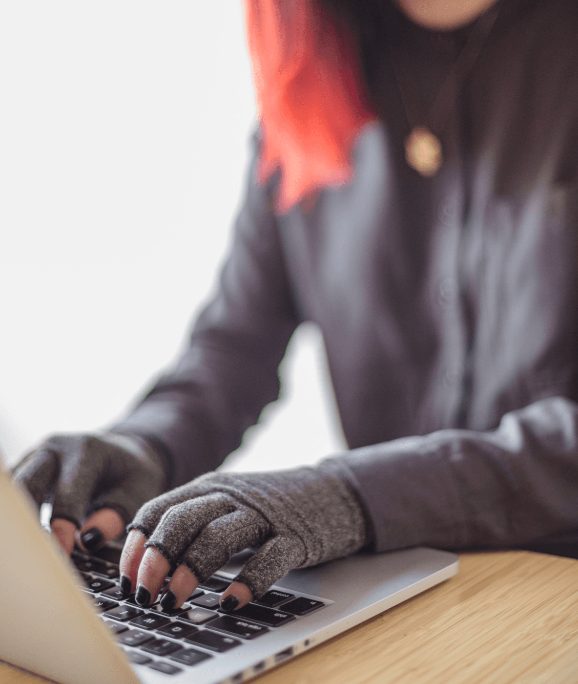 A disabled Asian genderfluid person types on a laptop while wearing compression gloves. The hands and keyboard are the focal point.