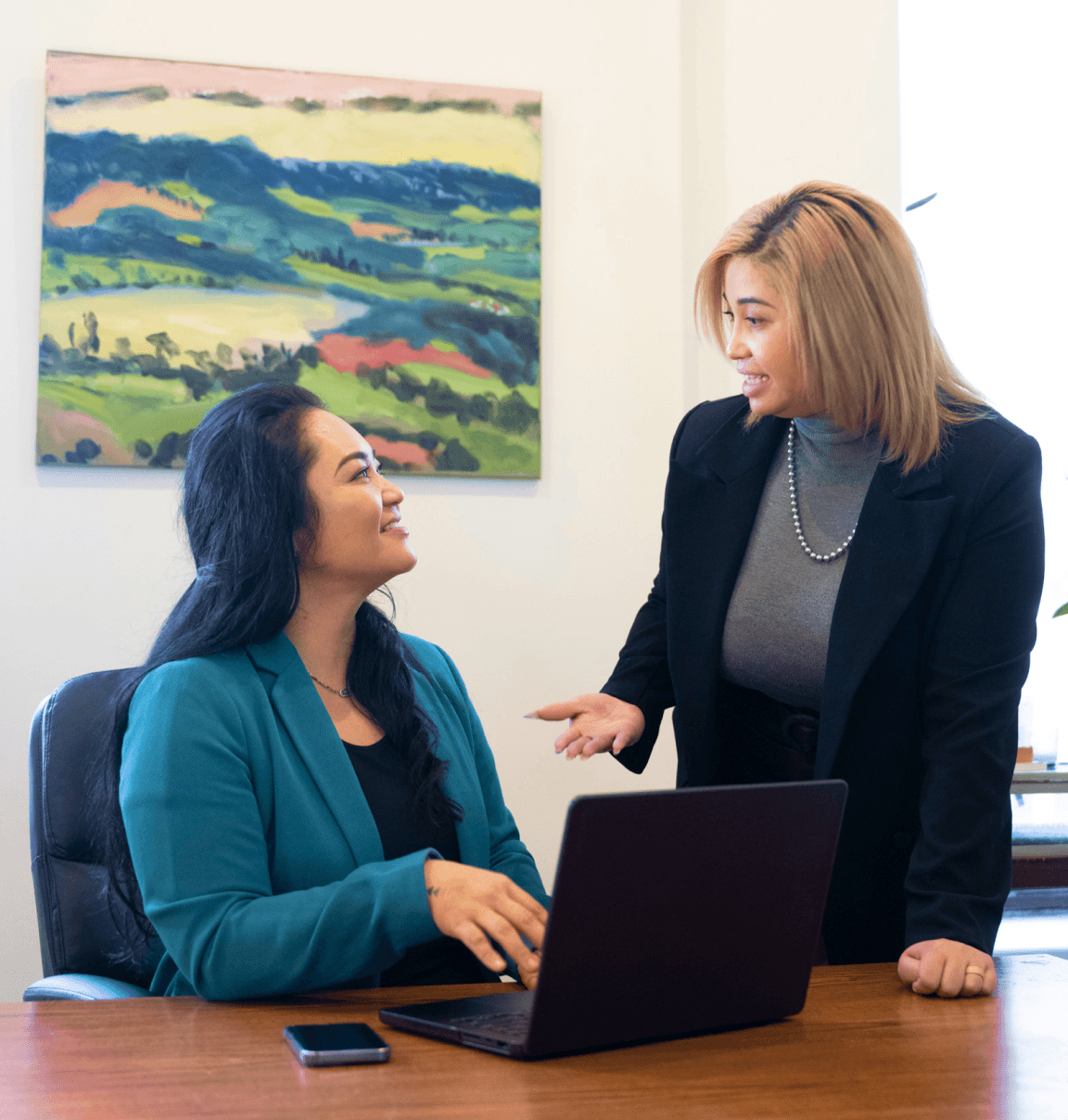 Two women at work speaking to one another in front of a desk with a laptop and mobile device on top of it.